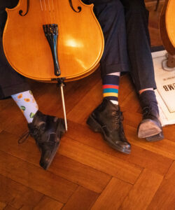 Das Bild zeigt zwei Paar Füße von Musiker:innen und ihre Musikinstrumente dazwischen. Dabei schauen bunte Socken hervor.