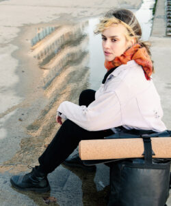 Auf dem Bild sieht man wie Phoebe von Yogoja an der Promenade des Spreeufers in Berlin sitzt. Neben ihr befindet sich ihre Yogamatte aus Kork, zusammen gerollt am Rucksack befestigt.